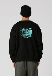 Persona con un gorro negro y sudadera negra con un diseño abstracto en turquesa en la espalda, de pie contra un fondo claro y uniforme.