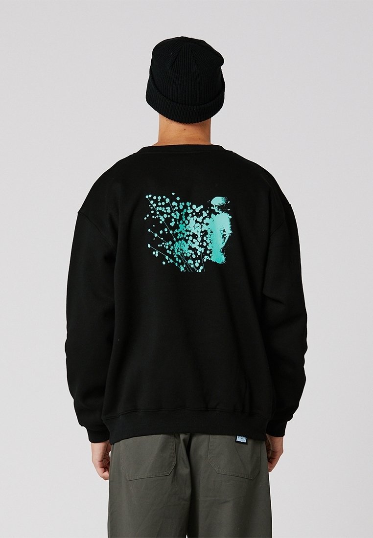 Persona con un gorro negro y sudadera negra con un diseño abstracto en turquesa en la espalda, de pie contra un fondo claro y uniforme.
