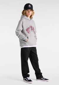 Sudadera gris con diseño gráfico rojo, bolsillos frontales, superpuesta a una camiseta blanca. Pantalones negros y zapatillas en blanco y negro completan el atuendo.