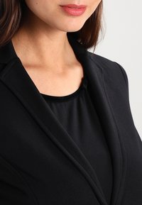 Blazer nero con una texture liscia, caratterizzato da revers a punta e un design aderente, indossato sopra un top nero a scollo ampio.