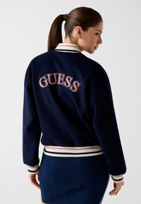 Blouson aviateur bleu marine en tissu doux, avec "GUESS" brodé en rose dans le dos, et des poignets et ourlet côtelés à rayures.