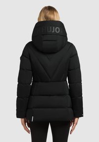 Žena v černé zateplené bundě s vysokým límcem a kapucí, na zádech s nápisem značky "KHUJO".