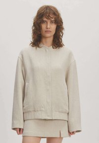 Helle beige, übergroße Jacke aus strukturiertem Stoff mit einer Knopfleiste, langen Ärmeln und seitlichen Taschen. Der Kragen ist rund und hat keine Kapuze.