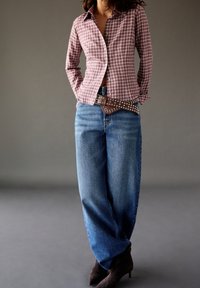 Czerwono-brązowa koszula w kratę o dopasowanym kroju, zestawiona z luźnymi niebieskimi dżinsami i skórzanym paskiem z ćwiekami w kolorze brązowym. Brązowe buty na obcasie dopełniają całość.