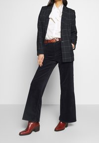 Czarna marynarka w kratę z usztywnionymi ramionami, biała bluzka z kokardą oraz granatowe rozszerzane spodnie sztruksowe. Brązowy pasek i czerwone botki za kostkę.