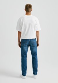 Camiseta blanca de algodón y vaqueros azul denim. La camiseta tiene un corte holgado; los vaqueros son rectos con bolsillos traseros y detalles de costura mínimos.