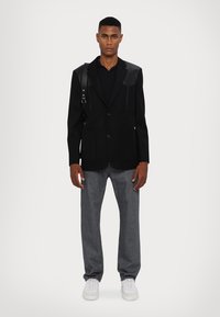 Zwarte blazer met leren schouders, gecombineerd met een zwart shirt, grijze jeans en witte sneakers. Voorzien van voorzakken en een getailleerd ontwerp.