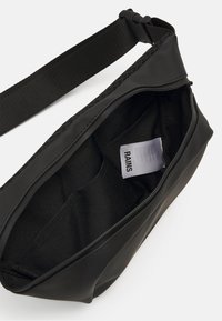 Juodas vandeniui atsparus krepšys, pagamintas iš lygaus medžiagos su užtrauktuku, turintis struktūruotą diržą ir audinį viduje su etikete.