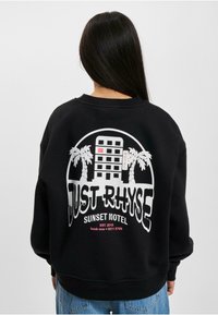 Fekete kapucnis pulóver, hátul nagy grafikával, amelyen a "JUST RHYSE" felirat, egy motel illusztráció és pálmafák fehér és rózsaszín színekben láthatók.