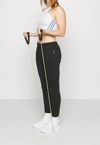 Svarta joggers med siddragkedjor, vit sportbh med tre ränder, håller i motståndsband, bär vita sneakers och crew-sockor.