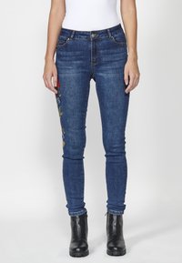 Blue denim skinny jeans with floral embroidery on the left leg, five pockets, and gold hardware details. Worn with black ankle boots.