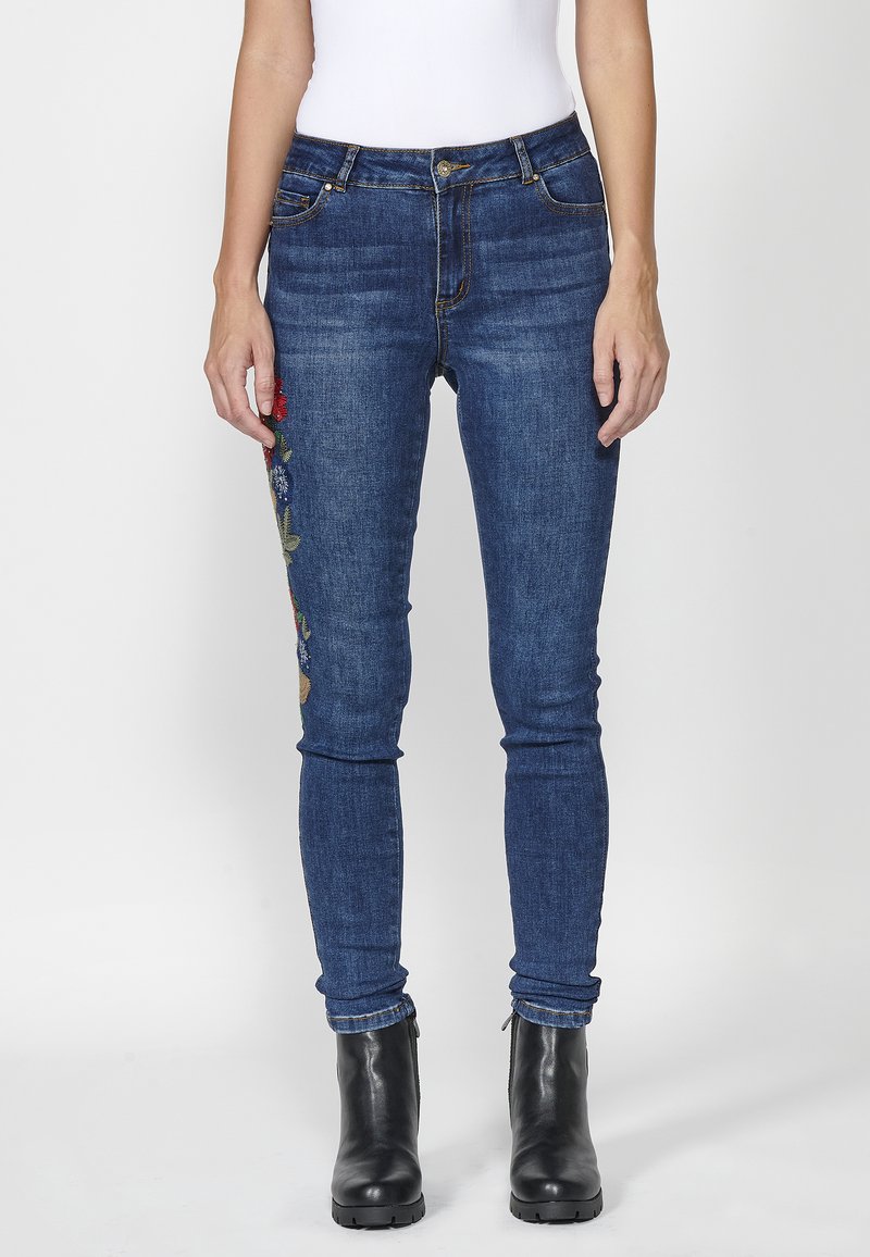 Blue denim skinny jeans with floral embroidery on the left leg, five pockets, and gold hardware details. Worn with black ankle boots.