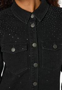 Detailní záběr na černou denimovou bundu s kovovými knoflíky, dvěma náprsními kapsami a malými rozptýlenými lesklými hroty na látce.