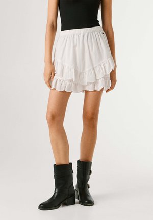 Vrouw in gelaagde witte rucheshort en zwarte leren laarzen tot halverwege de kuit, staand tegen een effen achtergrond.