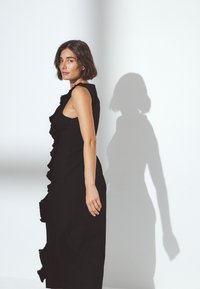 Musta mekko, jossa on röyhelöreunus sivuilla, hihaton malli, vartalonmyötäinen siluetti ja kuvioitu kangas. Varjo näkyy seinällä.