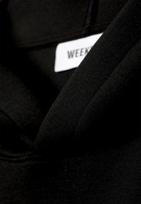 Sort hættetrøje med tekstureret stof, en synlig mærkat med teksten "WEEKEND" og en blød, let løs halsudskæring.