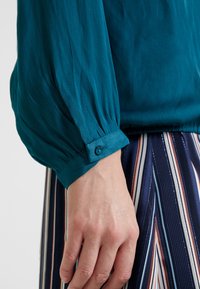 Gros plan d'une main près du poignet d'un chemisier à manches longues couleur sarcelle avec un bouton, au-dessus d'un pantalon rayé verticalement bleu marine, blanc et orange.