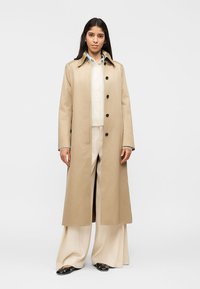 Γυναίκα που στέκεται φορώντας μπεζ μακρύ παλτό πάνω από λευκή ζακέτα και φαρδιά κρεμ παντελόνια με μαύρα παπούτσια τύπου slip-on.