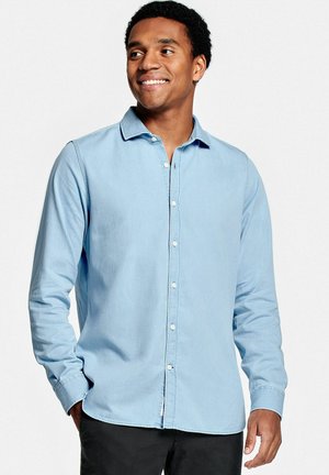 Man met krullend haar die glimlacht, gekleed in een lichtblauw overhemd met knopen en zwarte broek, staand met één hand in de zak tegen een witte achtergrond.