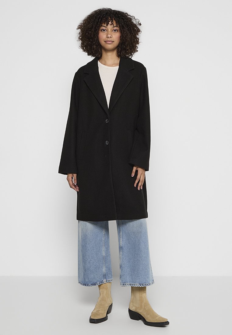 Zwarte wollen coat met een geknipte kraag, twee knopen, zijzakken en een losse pasvorm, gecombineerd met lichtblauwe wijde spijkerbroek en beige laarzen tot de enkel.