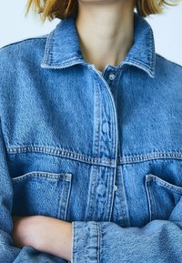 Světlomodrá džínová bunda s límcem, předními kapsami a zapínáním na knoflíky; obsahuje viditelné švy a volný střih.