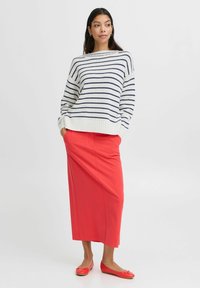 Gestreifter Strickpullover in Weiß und Marineblau, kombiniert mit einem korallenfarbigen Bleistiftrock und roten Ballerinas. Das Modell steht mit den Händen in den Taschen.