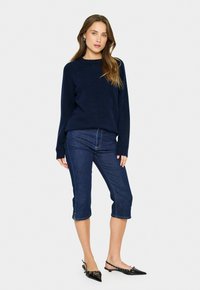 Ženska stoji v mornarsko modrem puloverju, temno modrih skrajšanih jeans hlačah in črnih usnjenih čevljih s koničastim nosom proti navadni beli ozadju.
