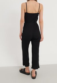 Mono negro de canalé con tirantes finos, cintura ajustada y piernas anchas. Presenta un diseño liso con una textura suave y un largo recortado.