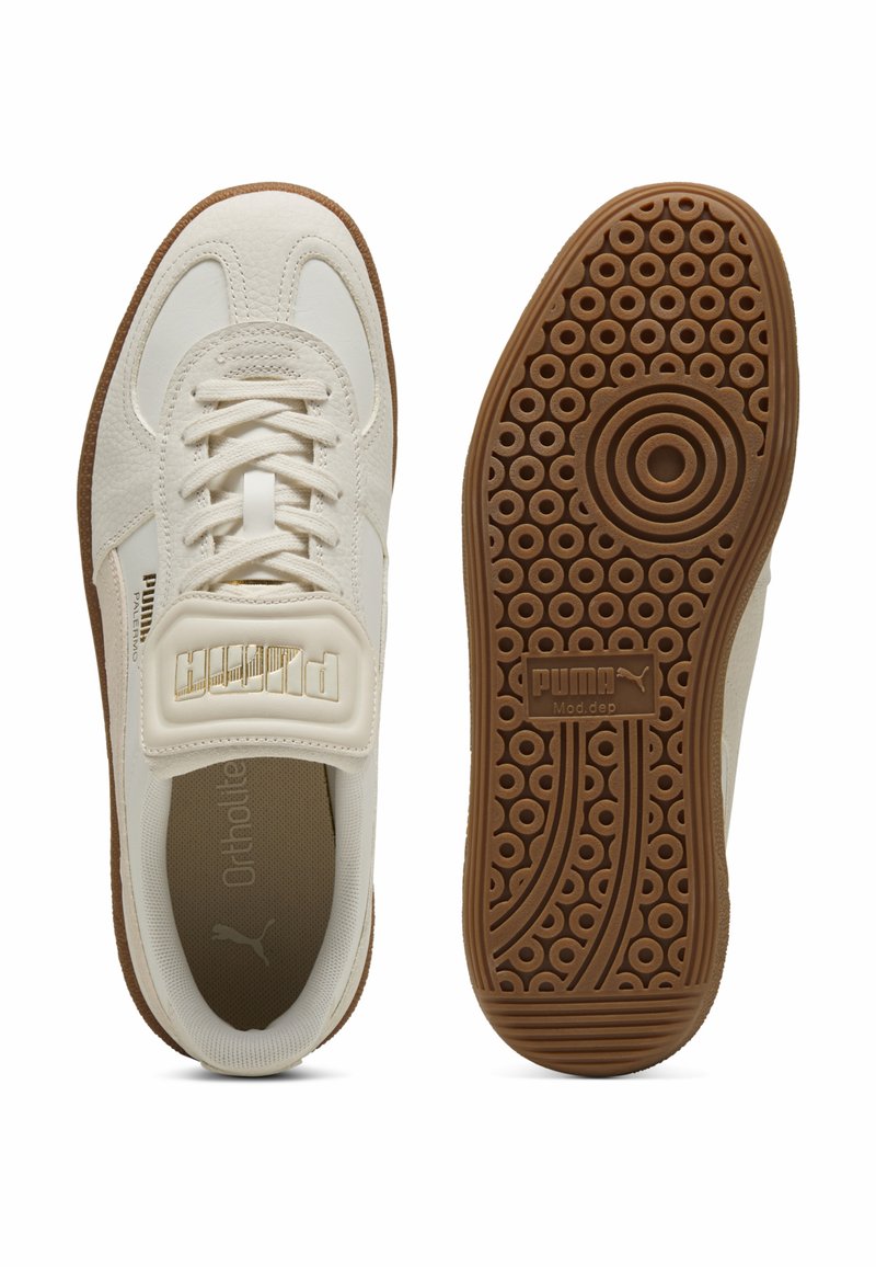 Baskets blanches avec un dessus en cuir texturé et une languette rembourrée, arborant un logo doré et une semelle en caoutchouc marron avec des motifs circulaires.
