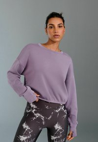 Heledad lilla lühike sweatshirt, mis on lõdva lõikega ja pikad varrukad, kombineeritud mustade marmorimustriga retuusidega. Sujuv tekstuur, minimaalne kaunistus.