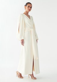 Vestido largo color crema con diseño cruzado, escote en V y mangas abullonadas. Cuenta con una abertura frontal y detalle de cintura atada. Tacones dorados con tiras.