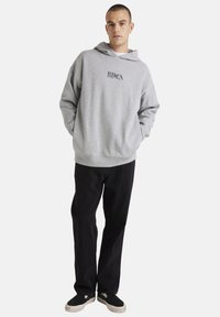 Grijze katoenen hoodie met een voorzak en een capuchon met trekkoord, voorzien van een zwart logo. Gecombineerd met zwarte broek en zwarte sneakers.