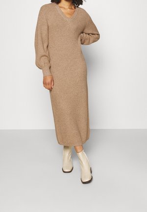 Abito a maglia con maniche lunghe e scollo a V in beige, caratterizzato da un motivo a rilievo e una silhouette rilassata, abbinato a stivali alla caviglia lucidi color crema.
