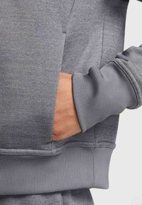 Main insérée dans la poche latérale d'un sweat-shirt gris avec poignets et taille côtelés visibles.