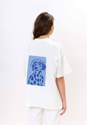 Witte katoenen t-shirt met een losse pasvorm, met een blauwe grafische afbeelding van een hond met vlekken en tekst: "Mijn therapeut heeft een natte neus!" op de achterkant.