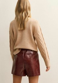 Beżowy sweter w splot kablowy o luźnym fasonie i teksturowanych rękawach, zestawiony z burgundowymi skórzanymi szortami o gładkim wykończeniu i wysokim stanem.