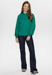 Camicia verde a maniche lunghe con colletto e polsini elastici, abbinata a jeans blu scuro e stivali beige, caratterizzata da un tessuto morbido e una vestibilità rilassata.