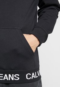 Ruka osoby v kapse černé mikiny s žebrovanými manžetami a pasem, která má na pasu bílé nápis "CALVIN JEANS".