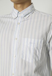 Homme portant une chemise à rayures verticales bleu clair et blanc, avec col boutonné et poche poitrine brodée du mot « GANT ».