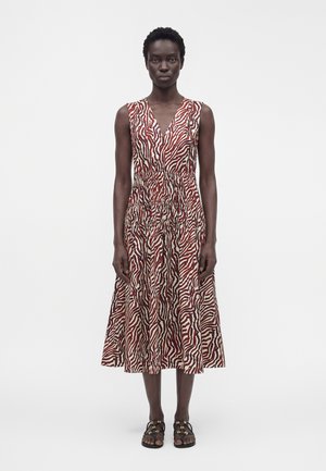 Femme debout, portant une robe midi sans manches à col en V avec un motif zèbre rouge et blanc et des sandales plates noires avec des détails dorés.