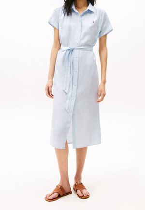 Vrouw draagt een lichtblauwe midi-jurk met korte mouwen en knopen, een gestrikte taille riem en bruine slip-on sandalen.