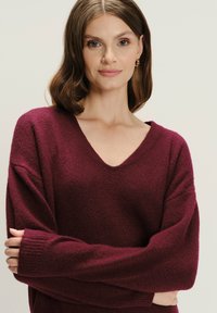 Bordeaux oversized sweater met een V-hals, geribbelde manchetten en een zachte textuur. Heeft brede mouwen en een ontspannen pasvorm, ideaal voor het laagjes dragen.