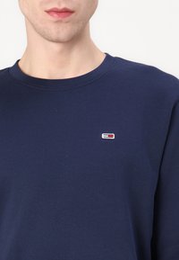 Man som bär en marinblå crewneck-tröja med en liten rektangulär röd, vit och blå logotyp på vänster bröst.