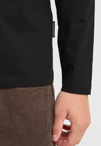 Čierna košeľa s dlhým rukávom a štítkom na boku, skombinovaná s hnedými textúrnymi nohavicami. Viditeľná ruka odpočívajúca na nohaviciach.