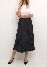 Czarna plisowana spódnica midi z elastycznym pasem, w zestawieniu z białym topem z krótkim rękawem i czarnymi szpilkami z ostrym czubkiem. Gładka faktura.
