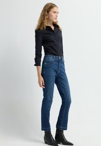 Jeans oscuros de corte ajustado y cintura alta, con diseño clásico de 5 bolsillos y costuras amarillas; combinados con una camisa negra de manga larga y botones.