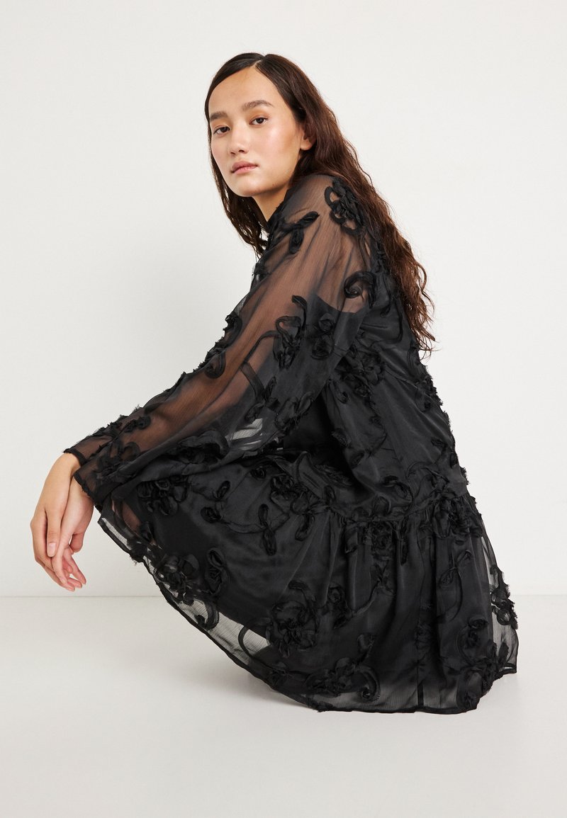 Crna prozirna haljina s cvjetnim vezom, dugim rukavima, naboranim strukom i lepršavim siluetom. Glatka tekstura i lagana tkanina.