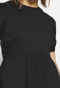 Femme portant une robe noire à manches courtes avec une taille froncée et des épaules bouffantes, sur un fond clair uni.