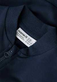 Tumši zils zip-up jakas ar ribotiem apkakli un piedurkņu galotnēm. Ietver zīmola aprūpes etiķeti ar izmēru un ražošanas informāciju.