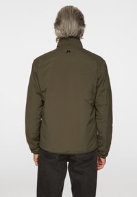 Olivgrön jacka med ståkrage, lättviktsmaterial och diskret logotypdetalj på baksidan. Kombinerad med mörka byxor.